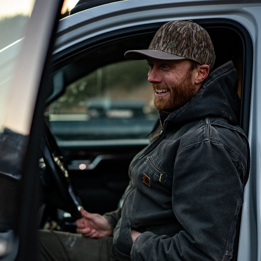 Man sitting inside a vehicle wearing a camouflage hat and dark jacket.