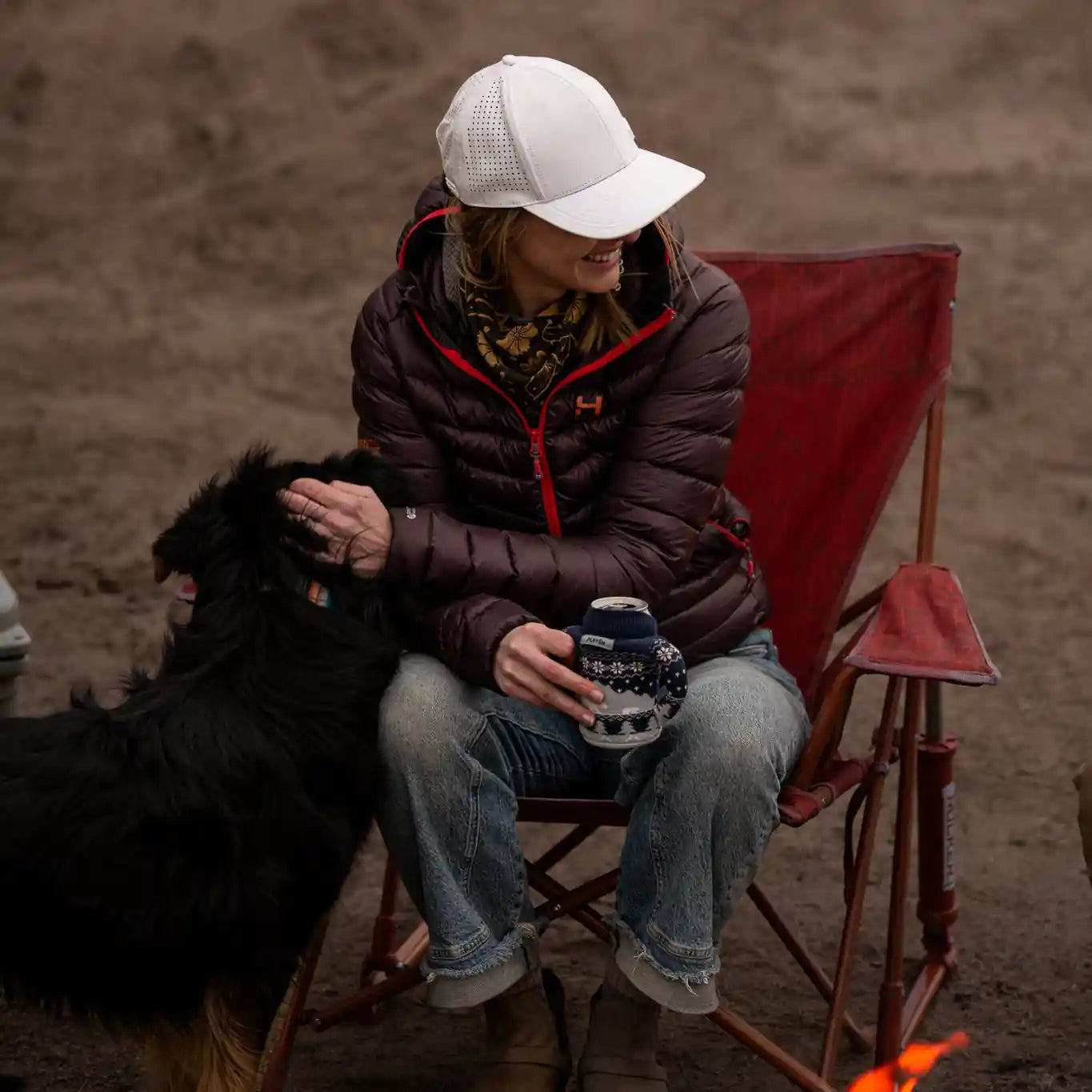 Person sitting by a fire with a dog, wearing a white cap and brown jacket.