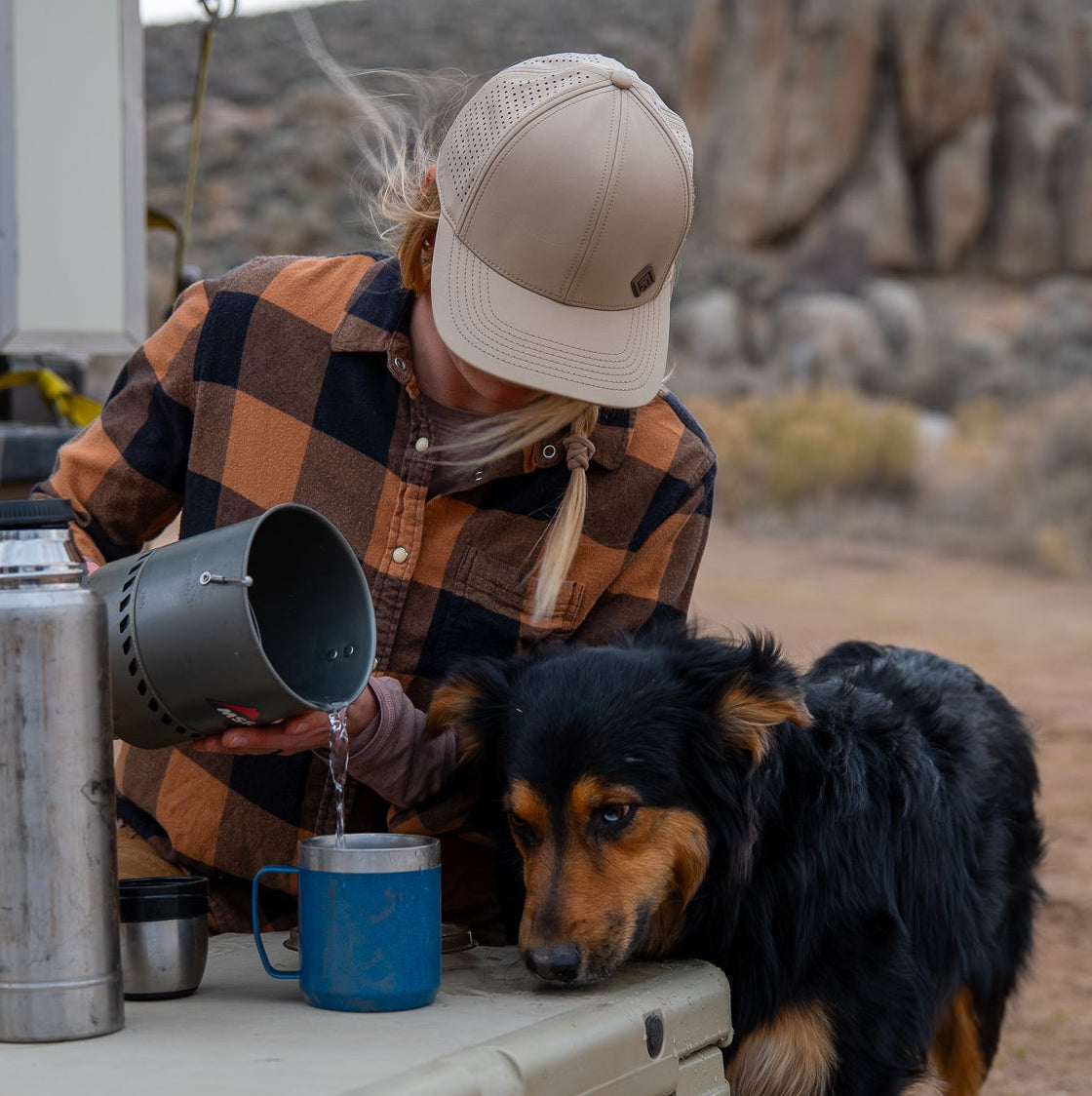 Person in plaid shirt and cap pouring from a container into a blue cup next to a dog outdoors.