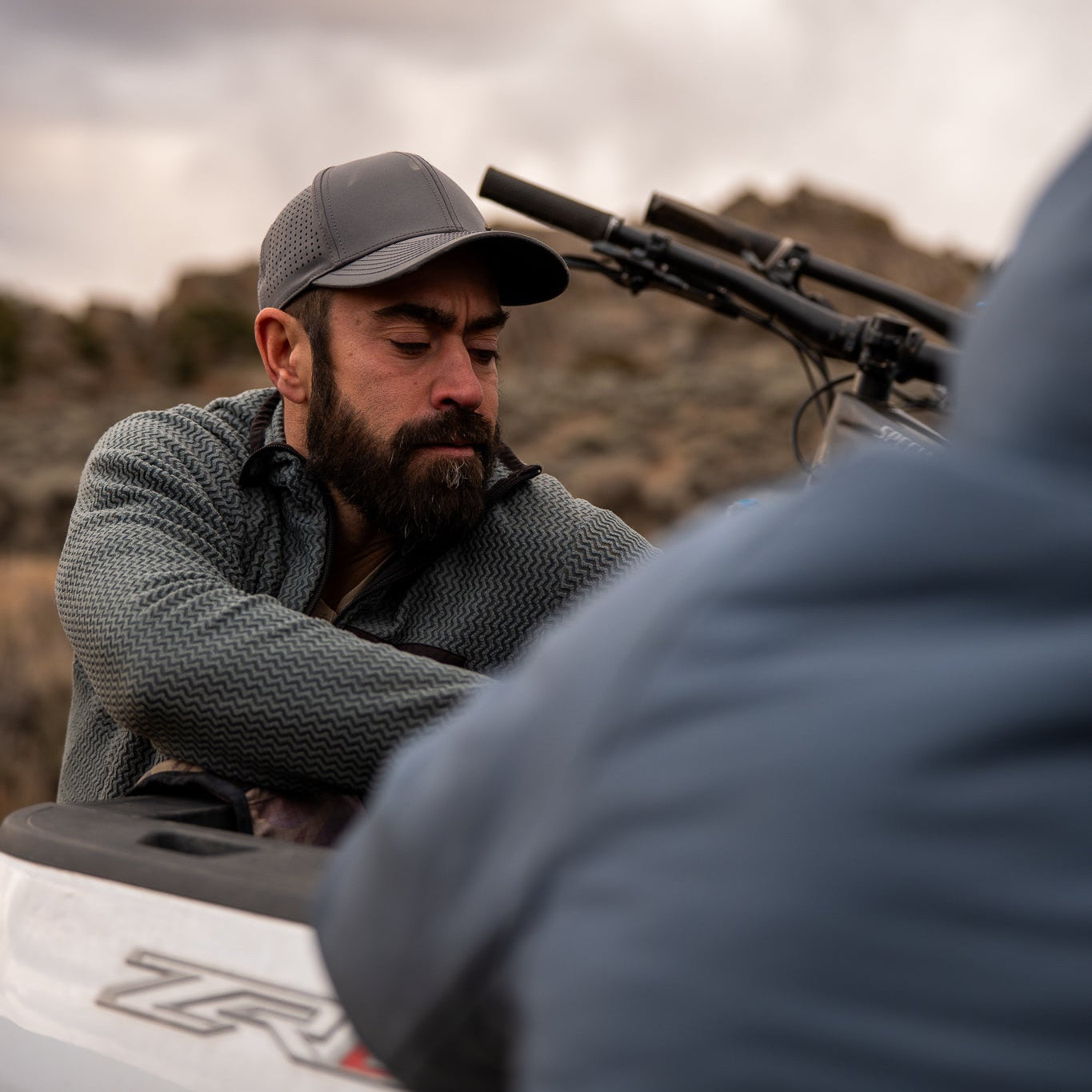 Man with a beard and cap interacting with another person outdoors, with a vehicle and rifle in the background.