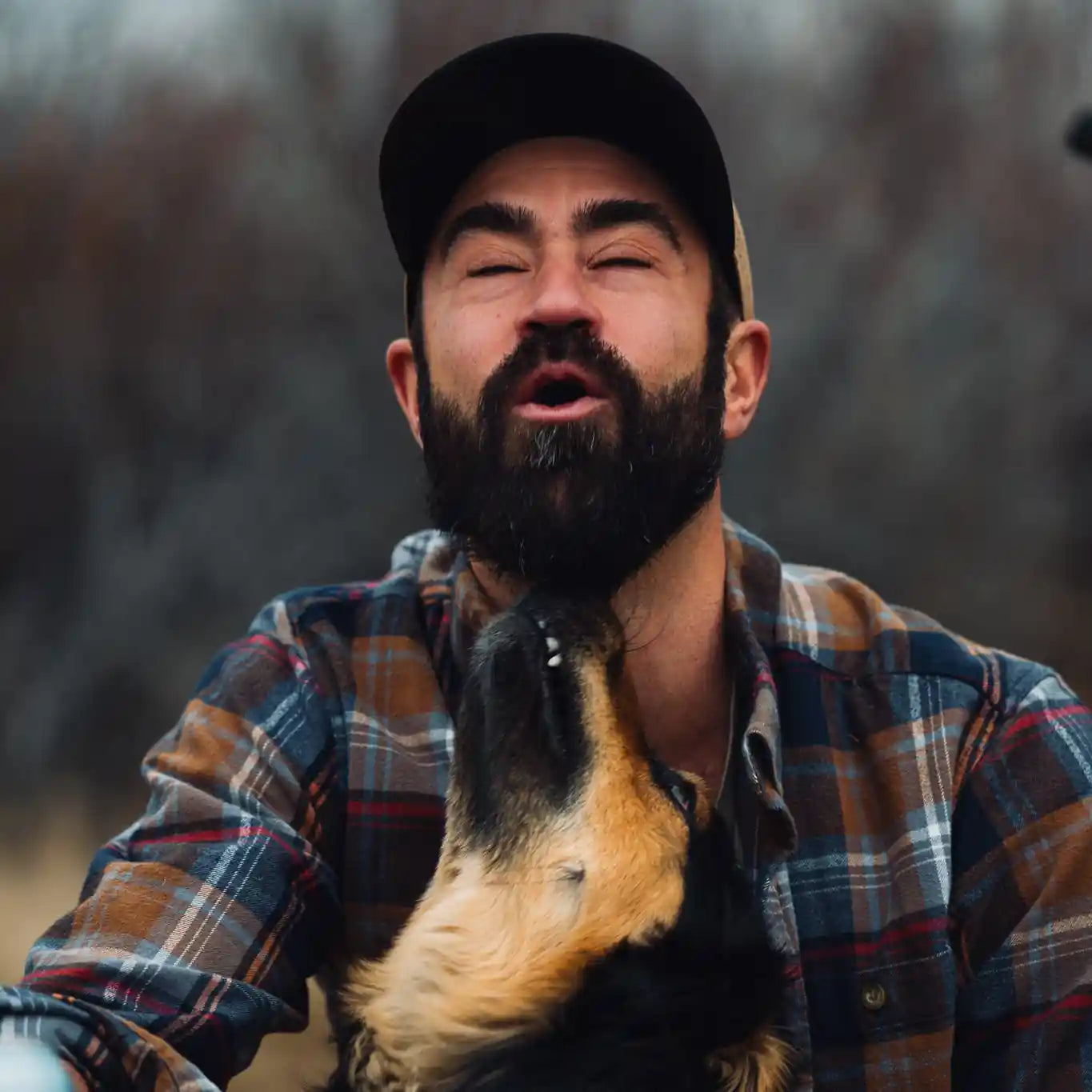 Man with a beard wearing a plaid shirt and cap, sitting outdoors with a dog on his lap.