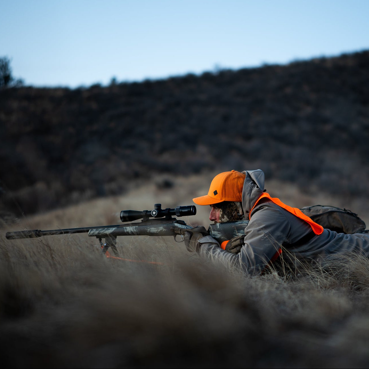 Hunter in orange vest and hat lying in grass with rifle, against a mountainous background