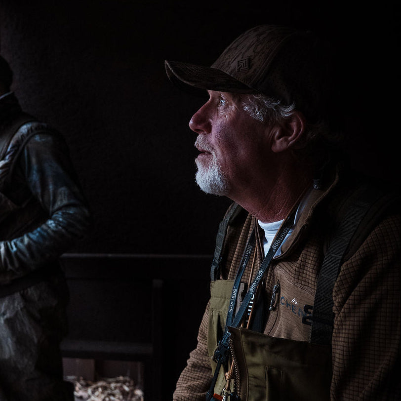 Two men in hunting attire inside a wooden cabin.