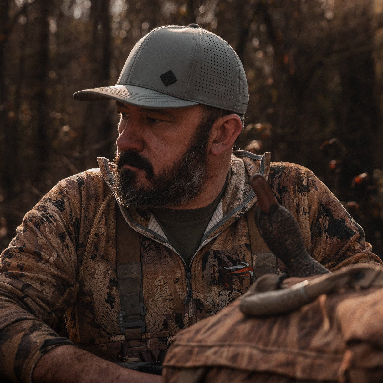 Man in camouflage clothing and cap sitting in a forest setting with a bag beside him.