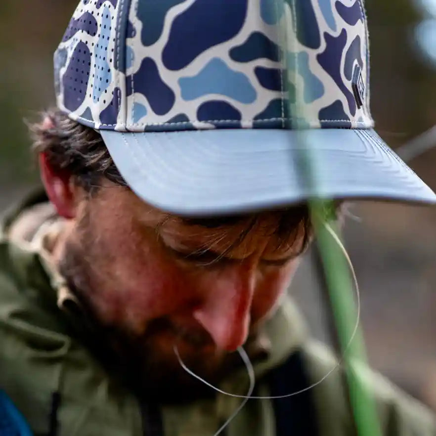Man wearing a blue camouflage cap and green jacket, holding a fishing rod.