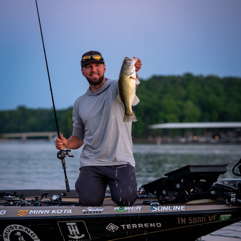 Man on a boat holding a large fish with fishing equipment, surrounded by water and trees.