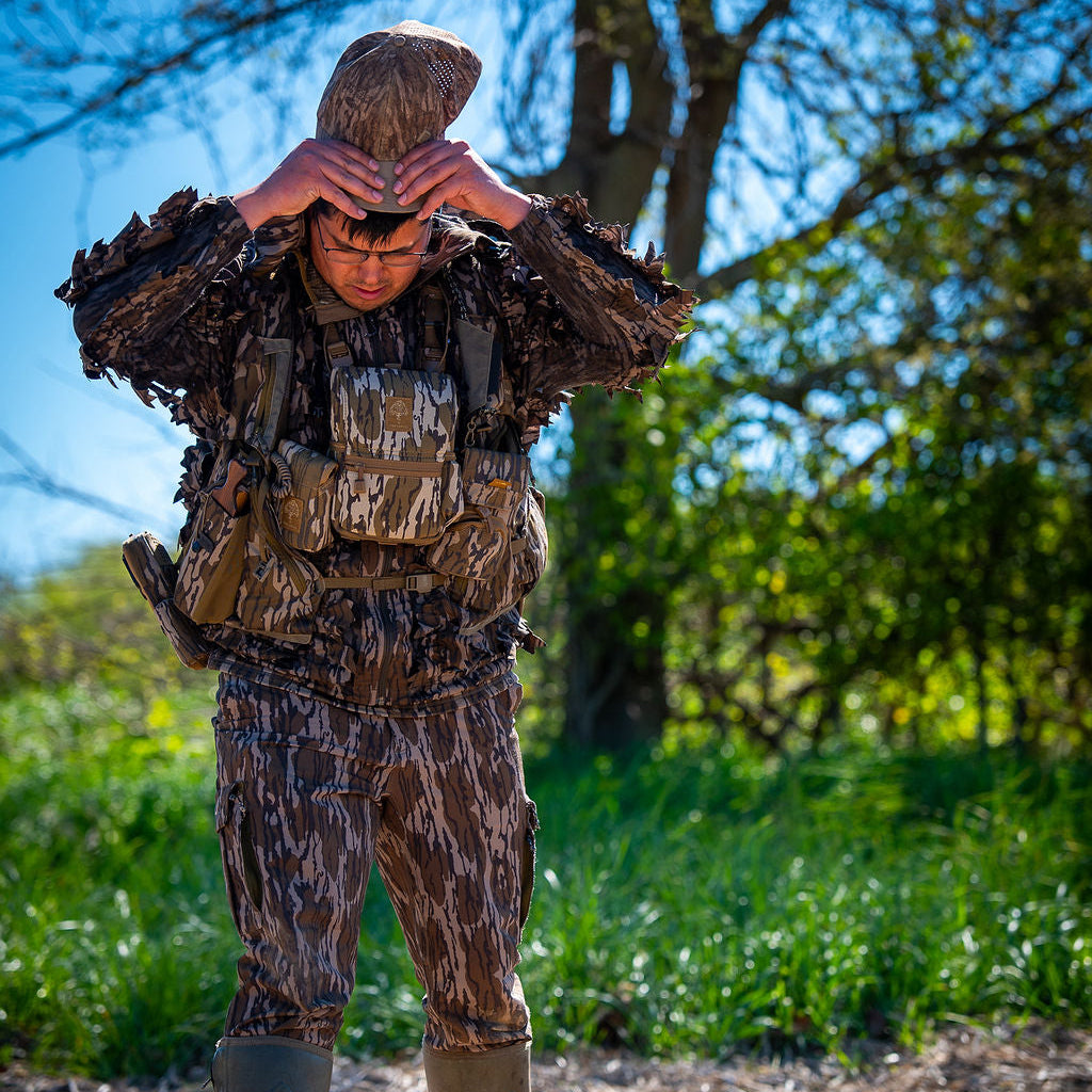 Person in full camouflage gear standing outdoors with trees and grass in the background