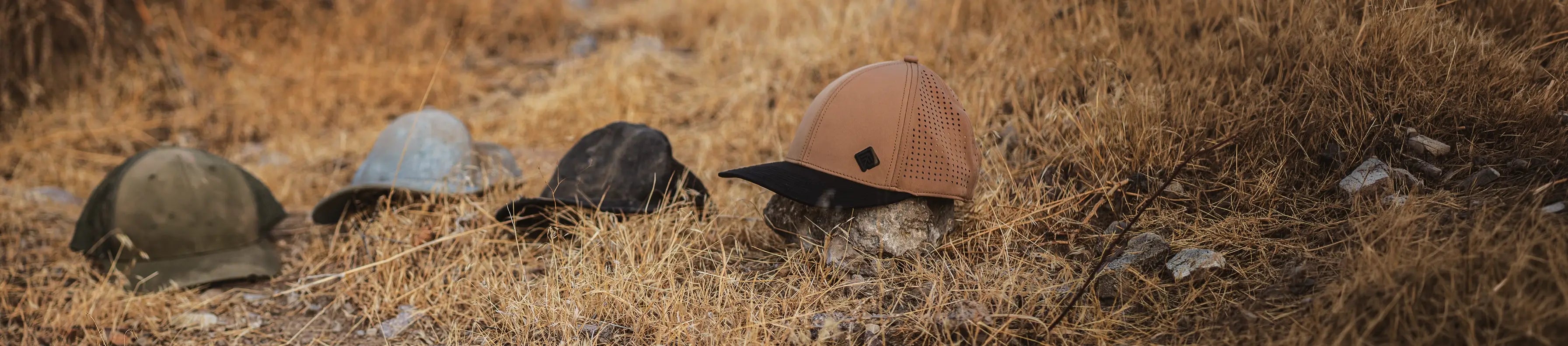 Three old tattered hats in various colors (olive green, light grey, black, and brown) and one new Terreno hat arranged in a row on dry, golden-brown grass outdoors.