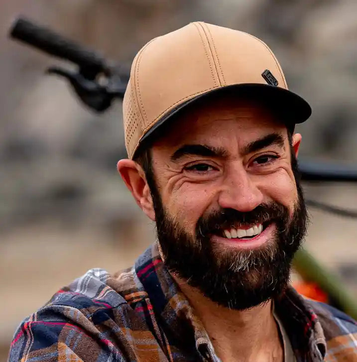 Man sitting outdoors with a bicycle, wearing a plaid shirt and cap.