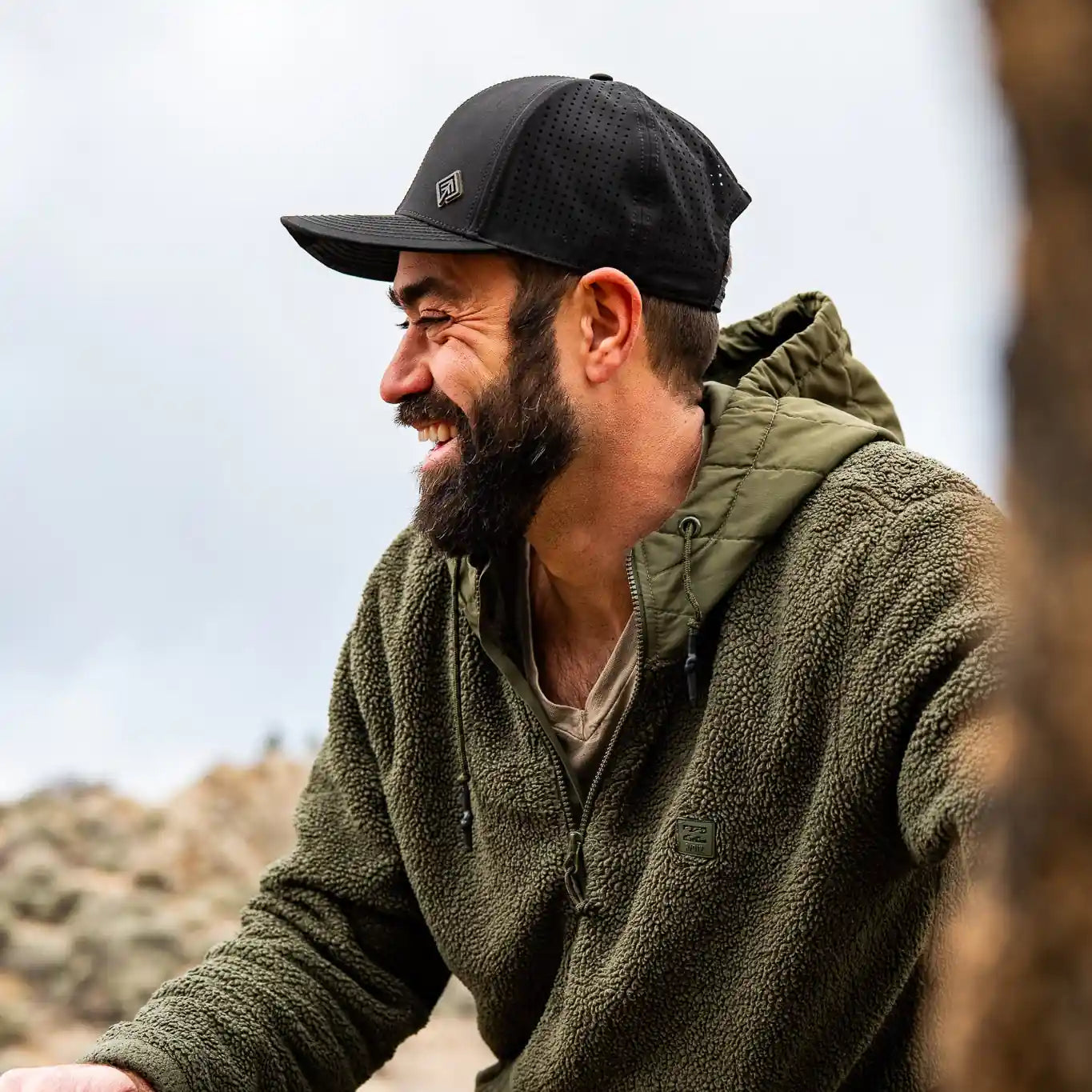 Man with a beard wearing a black cap and green jacket on a bike, with a blurred natural background