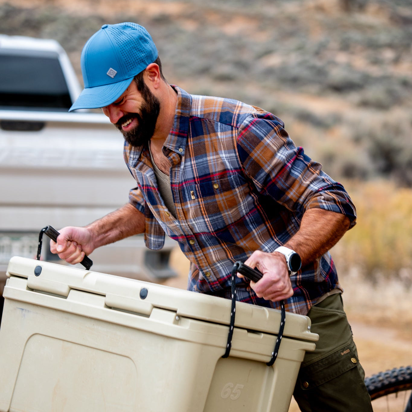 Man carrying a large beige cooler outdoors with a dog and bicycle in the background