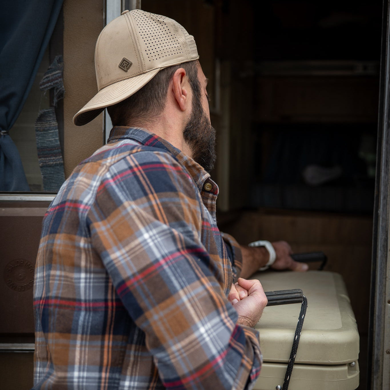 Man in plaid shirt and cap opening a cooler outdoors