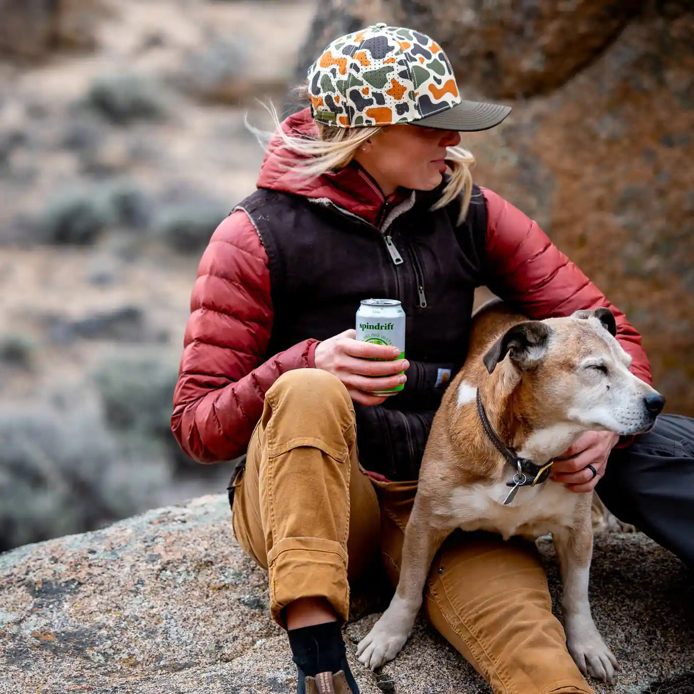 Person sitting on a rock with a dog, holding a can, in a natural outdoor setting.