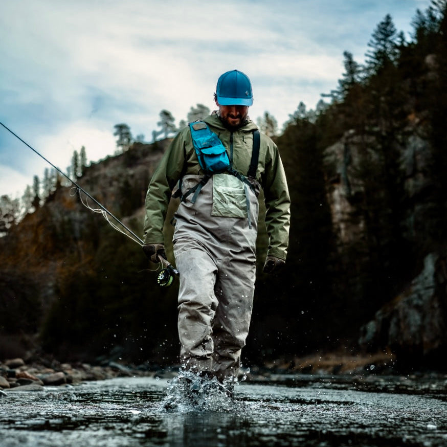 Person walking through a river with fishing gear, surrounded by nature.