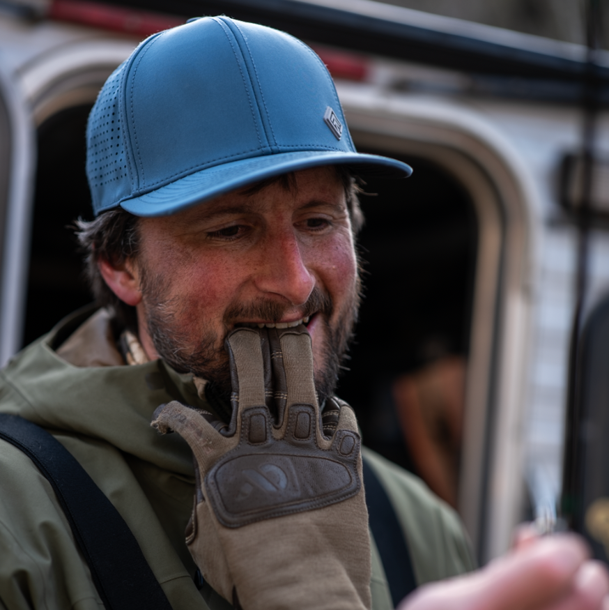 Man wearing a blue cap and brown gloves outdoors, possibly near a vehicle.