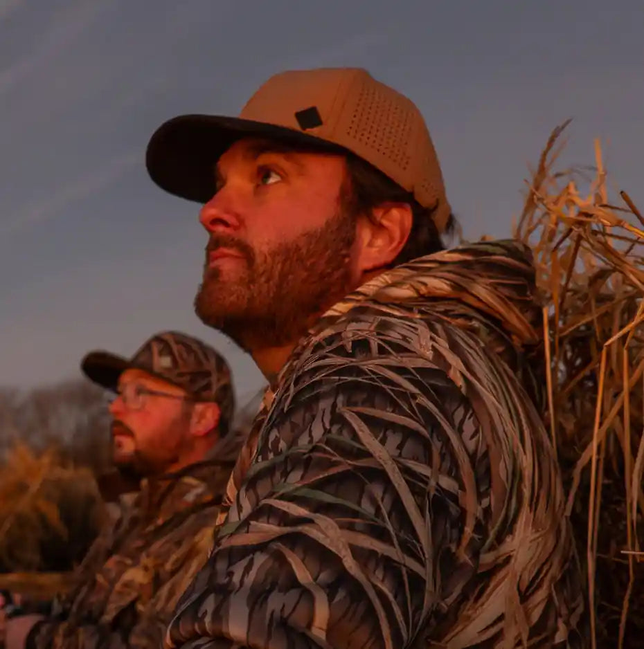 Two hunters in camouflage gear sitting in a field with a sunset sky.