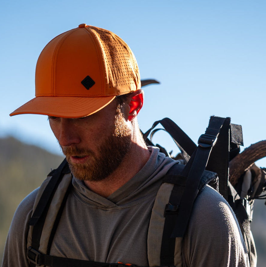 Man with orange cap and backpack holding rifle and deer antlers in a mountainous area