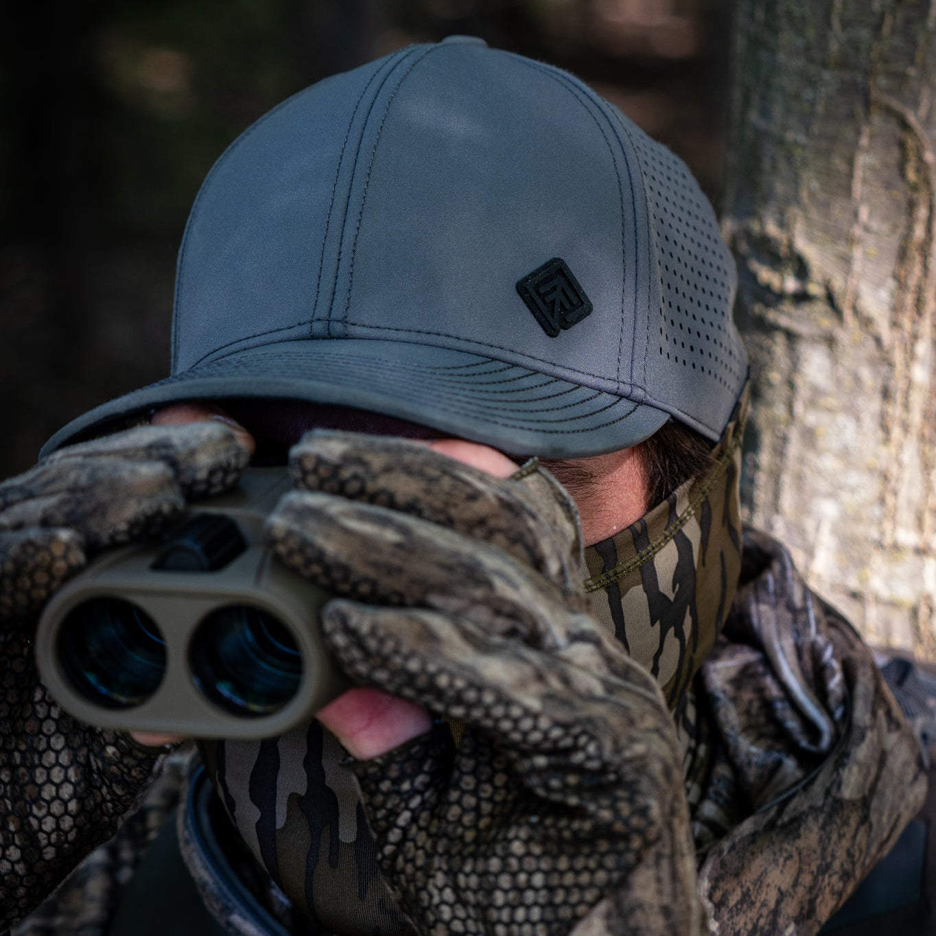 Person in camouflage gear using binoculars with a tree in the background