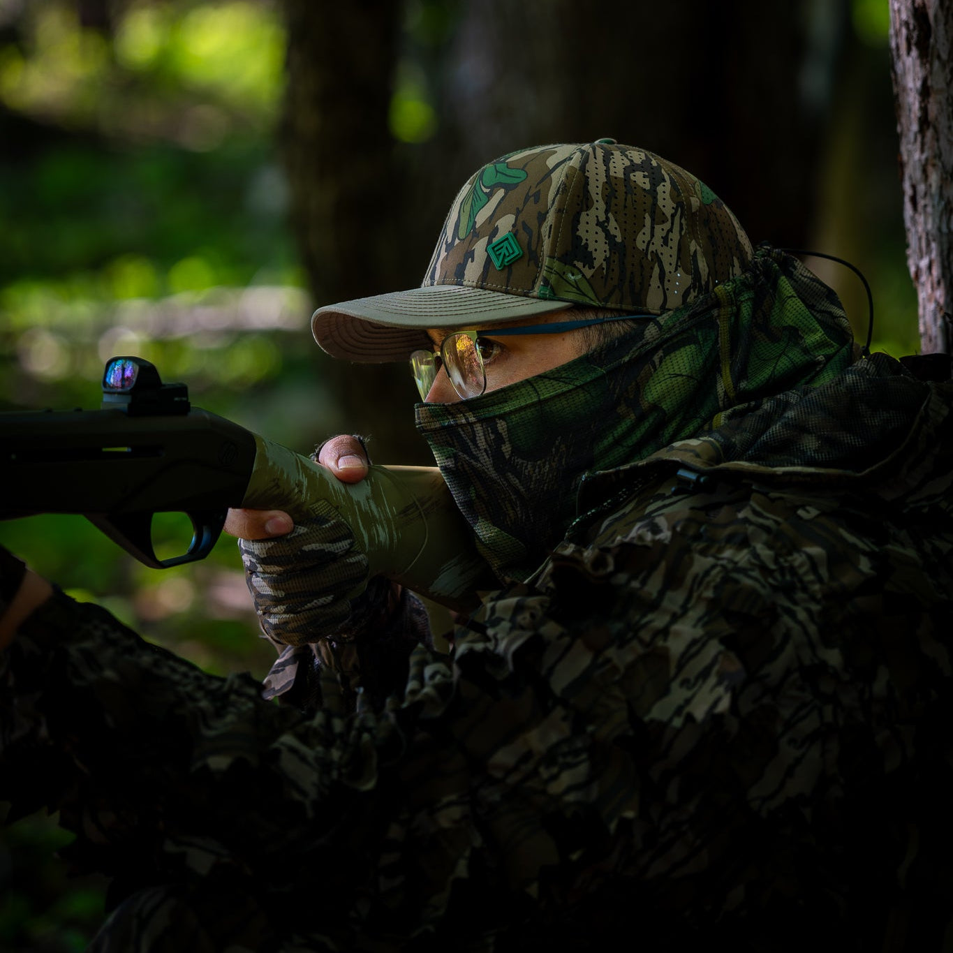 Person in camouflage gear with a rifle, peering through trees in a forest setting